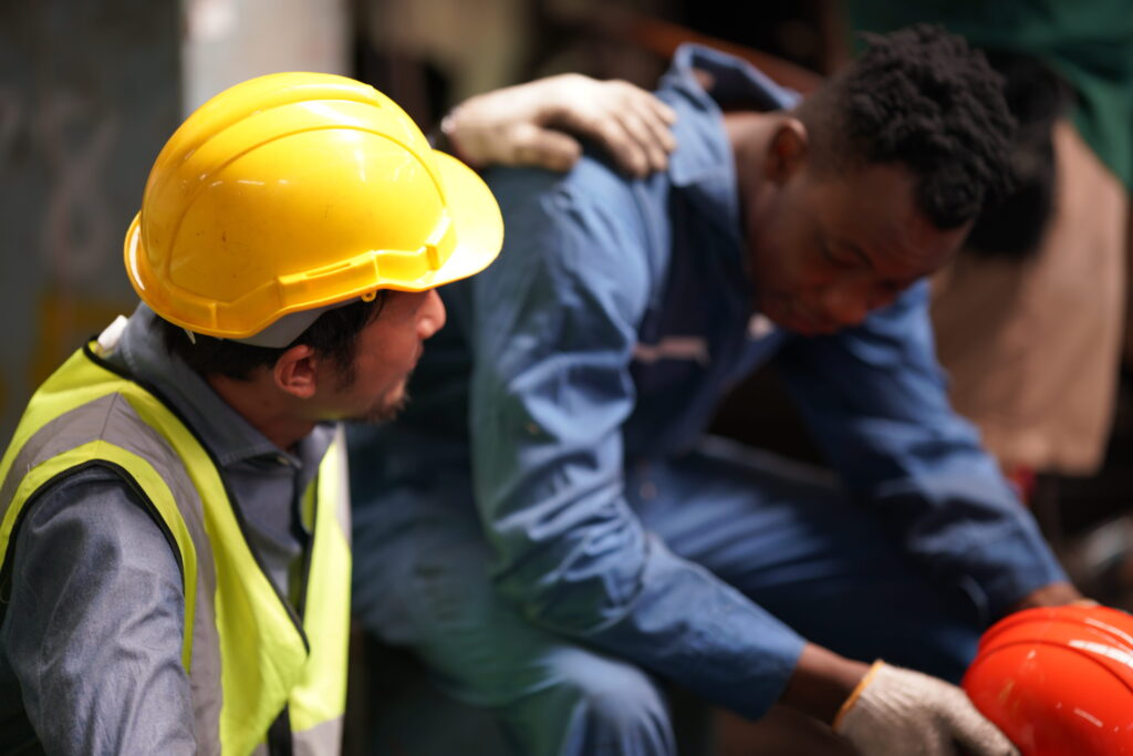 Construction worker in a yellow hard hat and safety vest checking on an injured coworker at a New Jersey job site