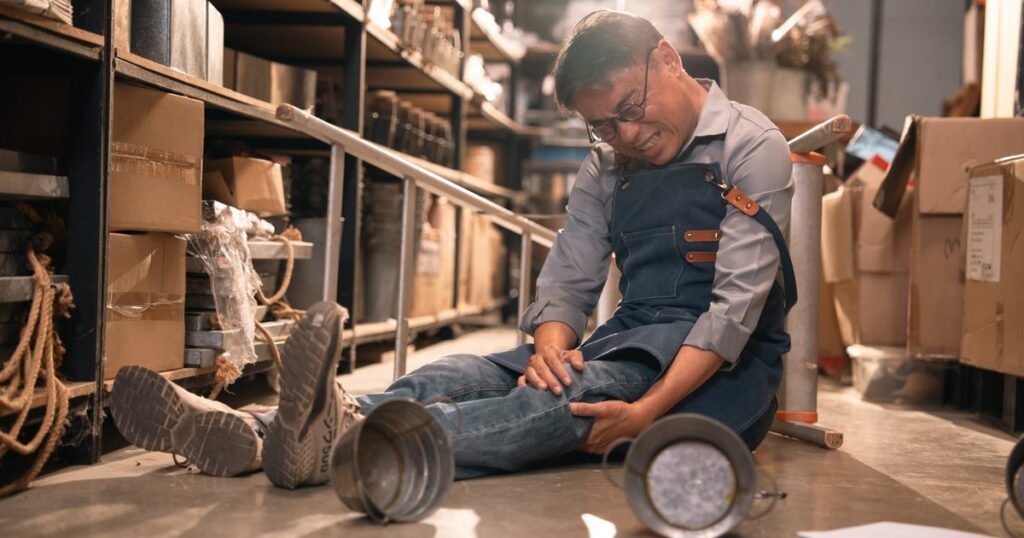 Warehouse worker sitting on the floor holding his injured knee after a fall, with stock shelves and a tipped stool behind him