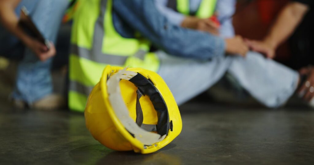 Yellow hard hat on the floor with an injured worker in a safety vest being helped in the background